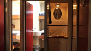 Een smalle gang die leidt naar een badkamer met een wastafel, spiegel en een deur met de tekst 'NOODUITGANG'.