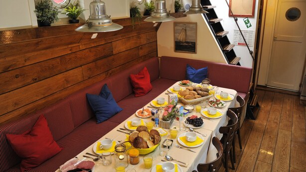 Een eetruimte met een lange tafel gedekt voor ontbijt, houten lambrisering aan de wanden en een bank met rode kussens.