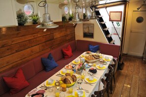 Un coin repas avec une longue table dressée pour le petit-déjeuner, des lambris en bois sur les murs et un banc avec des coussins rouges.