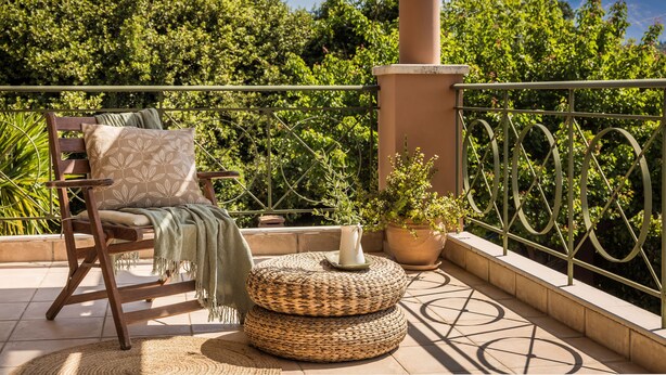 Ein Balkon mit einem Holzstuhl, einem gewebten Hocker, einem Blumentopf und einem Metallgeländer.