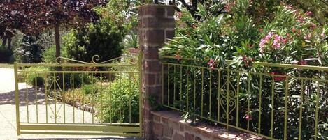 Une entrée fermée par un portail, avec un mur en briques et une clôture en métal, le tout entouré d’une végétation luxuriante et de plantes à fleurs.