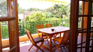 Ein offener Balkon mit einem Holztisch und Stühlen, Blick auf Grünflächen und Gebäude, und ein gelb gestreiftes Markisen