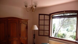 Ein Schlafzimmer mit Holzschrank, einem Bett mit rot-weiß gestreifter Bettdecke, einem grünen Sessel, einer Stehlampe und einem Fenster mit Blick auf Bäume.