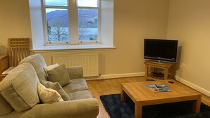 A living room with a sofa, a coffee table, a TV on a stand, and a window with a view of a landscape. - 3 bedroom 2nd floor apartment to sleeping 8 with stunning views of Loch Ness. (Loch Ness)