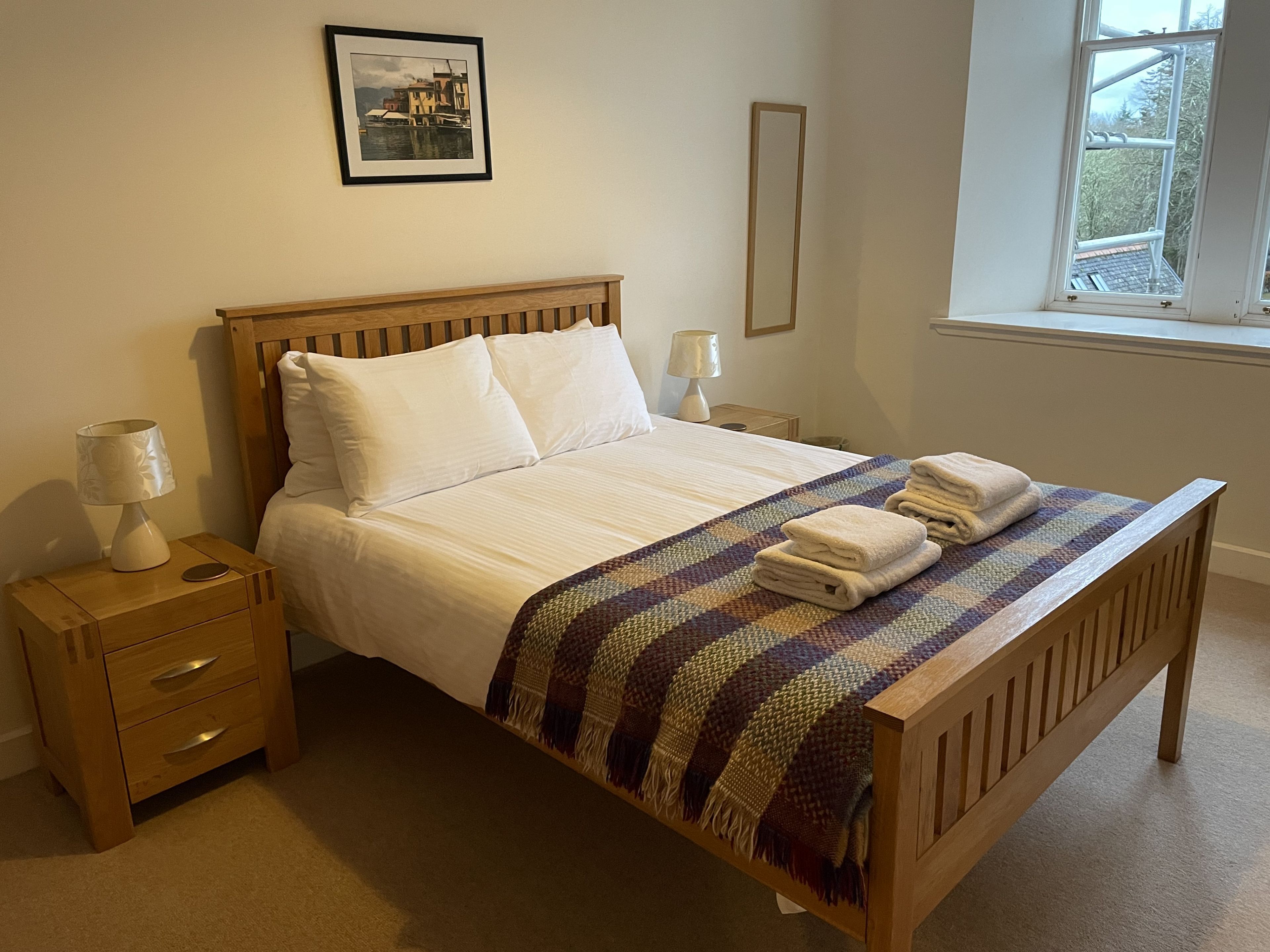 A neatly made bed with white linens, a wooden headboard, and a colorful woven blanket. There are two bedside tables with lamps and a framed picture on the wall.
