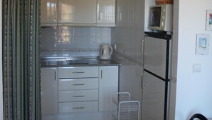 Een kleine keuken met witte kasten, een magnetron en een waterkoker. Er staat een klein wit krukje en een metalen rek.