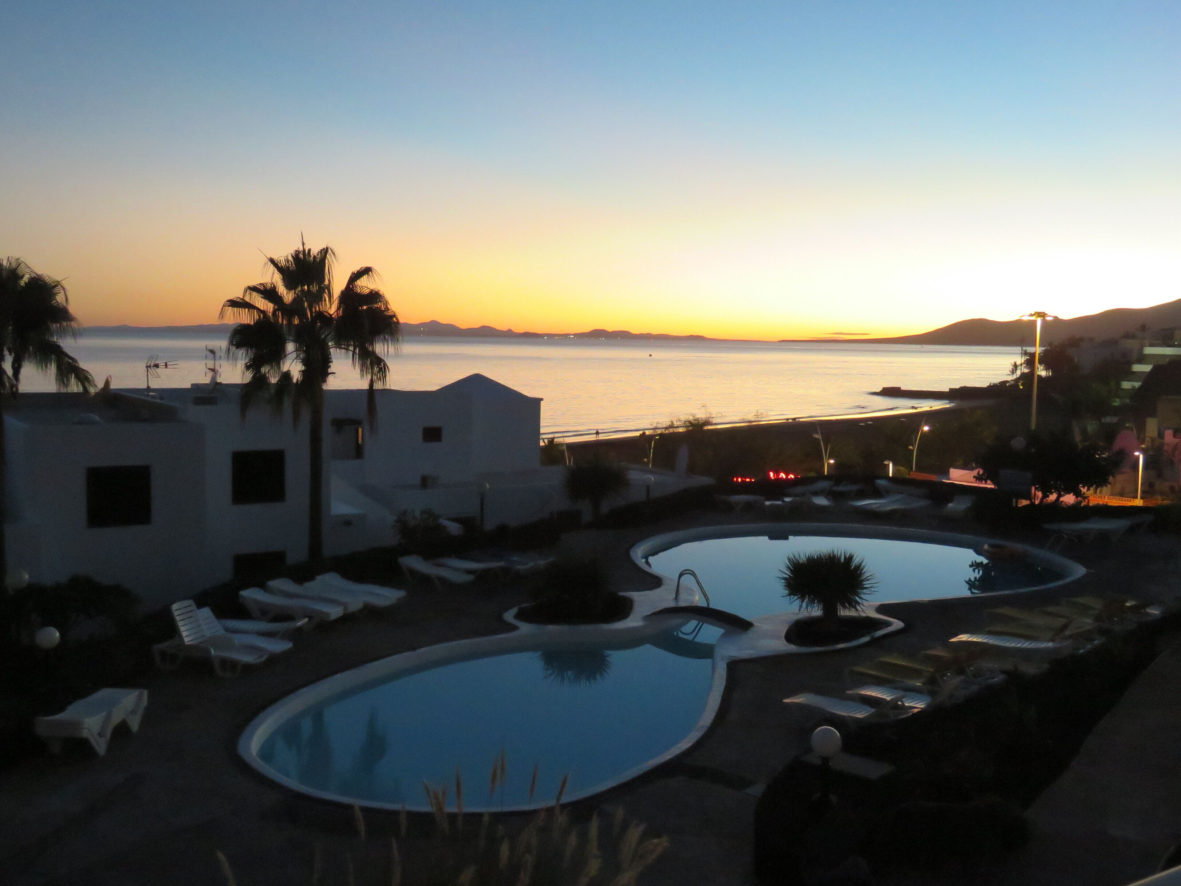 A resort pool area with lounge chairs, a swimming pool, and a view of the sea at sunset.