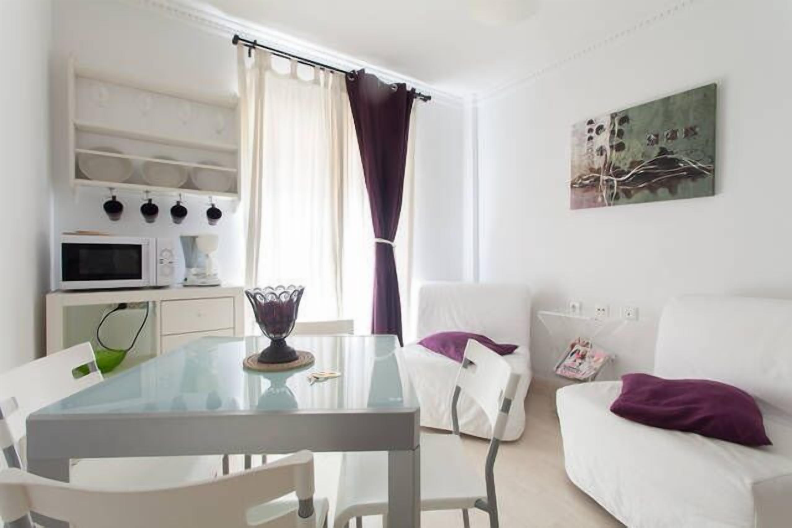 A modern living room with a glass dining table, white sofas, and a kitchen area with a microwave.