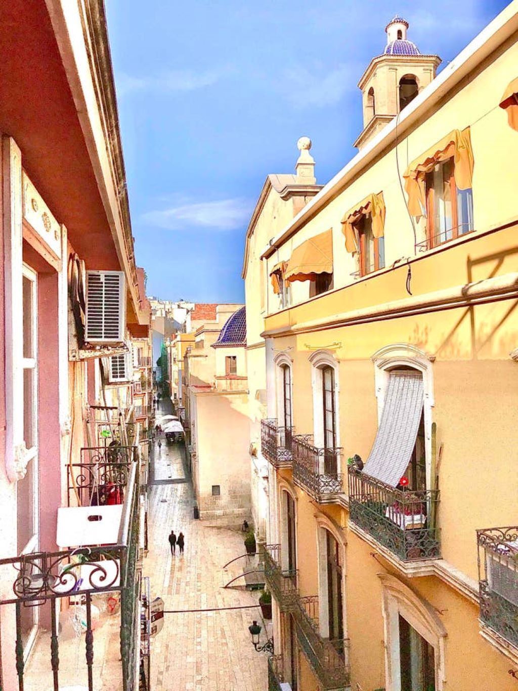 Une rue étroite, bordée de bâtiments des deux côtés, avec des balcons et un ciel dégagé.