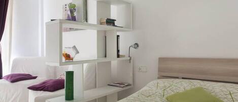 A modern bedroom with a bed, a white sofa, a built-in shelving unit, and a bedside table.