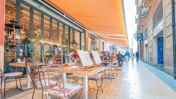 Een terras met tafels en stoelen, een zonnescherm, en een straatbeeld met gebouwen en voetgangers.