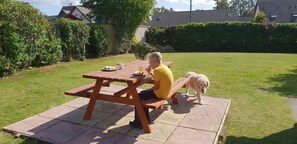 A person sitting at a wooden picnic table in a grassy backyard with a dog nearby.