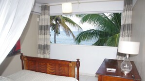 Quarto com cama de madeira, mesa de cabeceira com abajur e janela com vista para palmeiras e o mar.