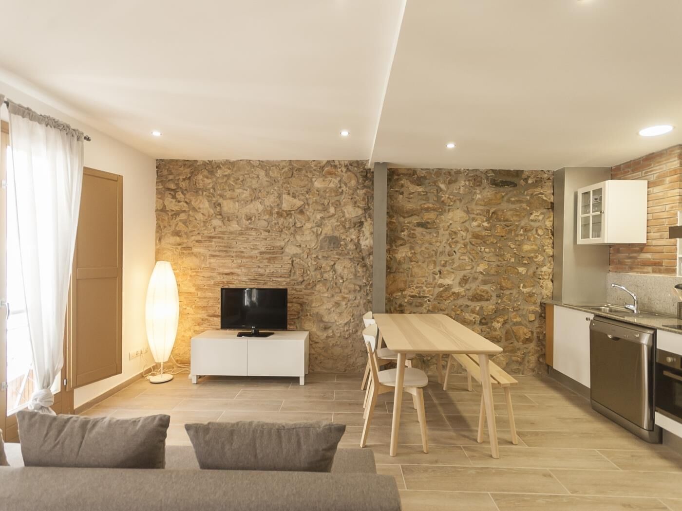 A modern living room with a stone wall, a flat-screen TV, a dining table with chairs, a sofa, and a kitchen area with a sink and appliances.