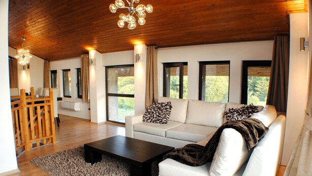 Ein großzügiges Wohnzimmer mit Holzbalkendecke, einer weißen Sofagarnitur, einem Couchtisch und großen Fenstern mit Blick auf Grünflächen.