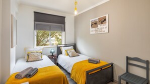 A bedroom with two beds, each with a yellow blanket and pillows, a nightstand with a lamp, a wooden chair, and a window with blinds. - Kwadalayo Art Quarters (Faro)