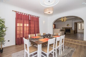Een eetruimte met een tafel voor zes personen, witte stoelen, een rood gordijn en een kroonluchter.