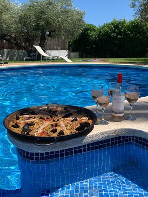 Ein Swimmingpool mit einem Paella-Gericht mit Meeresfrüchten und zwei Weingläsern am Rand.