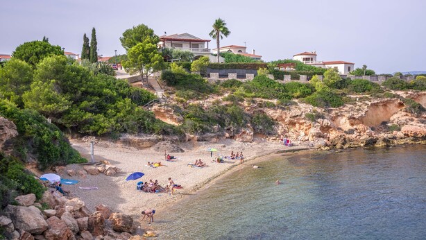 A coastal beach with clear waters, rocky terrain, and several buildings in the background.