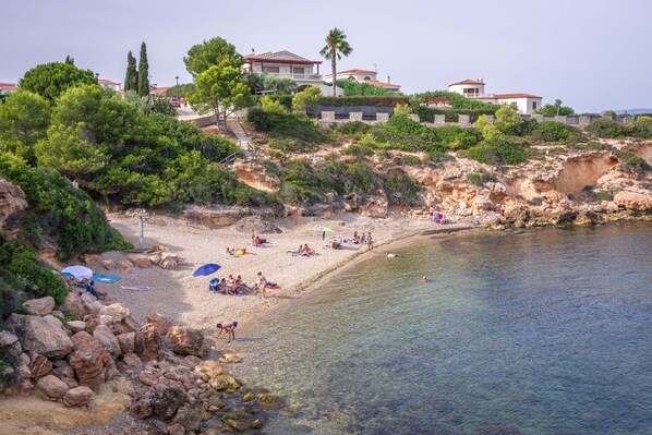 Ein Küstenstrand mit klarem Wasser, felsigem Gelände und mehreren Gebäuden im Hintergrund.