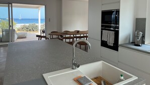 Una cocina moderna con una isla grande, horno empotrado y cafetera. Hay un comedor con mesa y sillas, y vistas al mar a través de una puerta abierta.