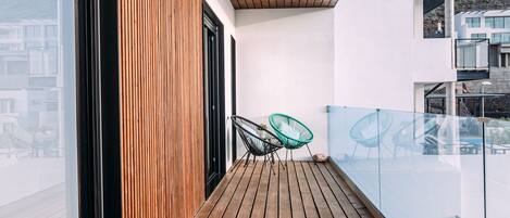 Une terrasse en bois avec une balustrade en verre, deux chaises vertes et une vue sur des bâtiments en arrière-plan.