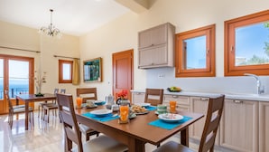 Une cuisine moderne avec des meubles en bois, une table à manger accompagnée de chaises, et une vue sur la mer grâce à de grandes fenêtres.