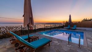 Un espace piscine avec des chaises longues et des parasols, un sol en pierre et une vue sur la mer au coucher du soleil.