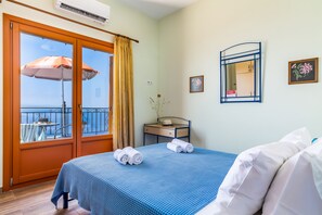 Une chambre d’hôtel avec un balcon, un lit avec une literie bleue, une table de chevet sur laquelle se trouve un chapeau, et un tableau encadré accroché au mur.