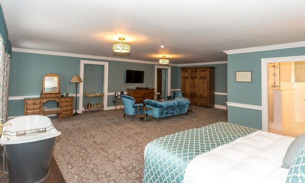 A hotel room with a large bed, a sitting area with a blue armchair, a vanity with a mirror, a wooden dresser, and a shower area.