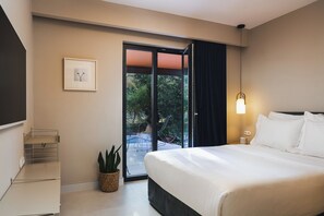 A hotel room with a bed, a small potted plant, a framed picture, a bedside table, and a window with a view of greenery. - Villa Elli - A magical garden retreat with private pools and curated spaces (Rethymno)
