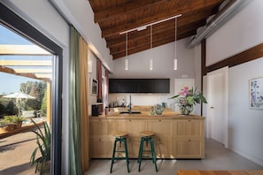A modern kitchen with wooden cabinets, a central island with stools, pendant lighting, and a view of the outdoors. - Villa Elli - A magical garden retreat with private pools and curated spaces (Rethymno)