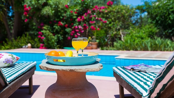 A poolside setting with a table holding oranges and a glass of orange juice, two lounge chairs, and a pool in the background.