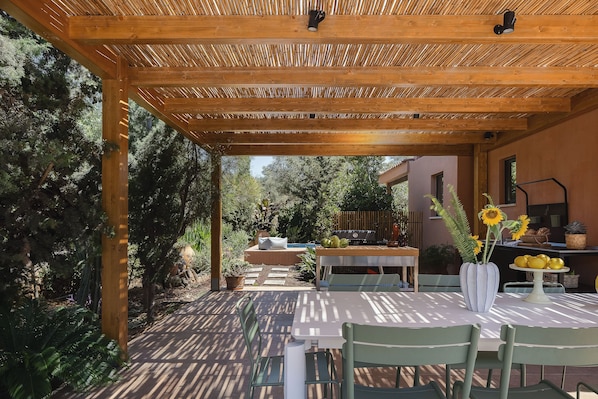 An outdoor patio with a wooden pergola, a table with chairs, and a variety of plants and fruits.