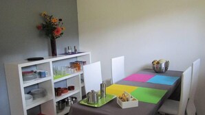 Un comedor con una mesa dispuesta para dos, un estante blanco con vajilla y vasos, y un jarrón con flores en la pared.