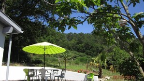 Un área de estar al aire libre con una mesa y sillas bajo un paraguas verde, rodeada de abundante vegetación y un edificio blanco.