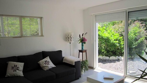Una sala de estar moderna con un sofá de color oscuro, una mesita auxiliar con un jarrón de flores y un amplio ventanal corredero que da a un jardín exterior.