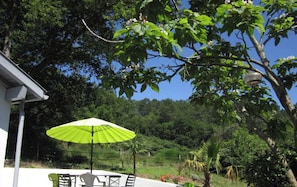 Een terras met een tafel en stoelen onder een groene parasol, omgeven door weelderige beplanting en een wit gebouw.
