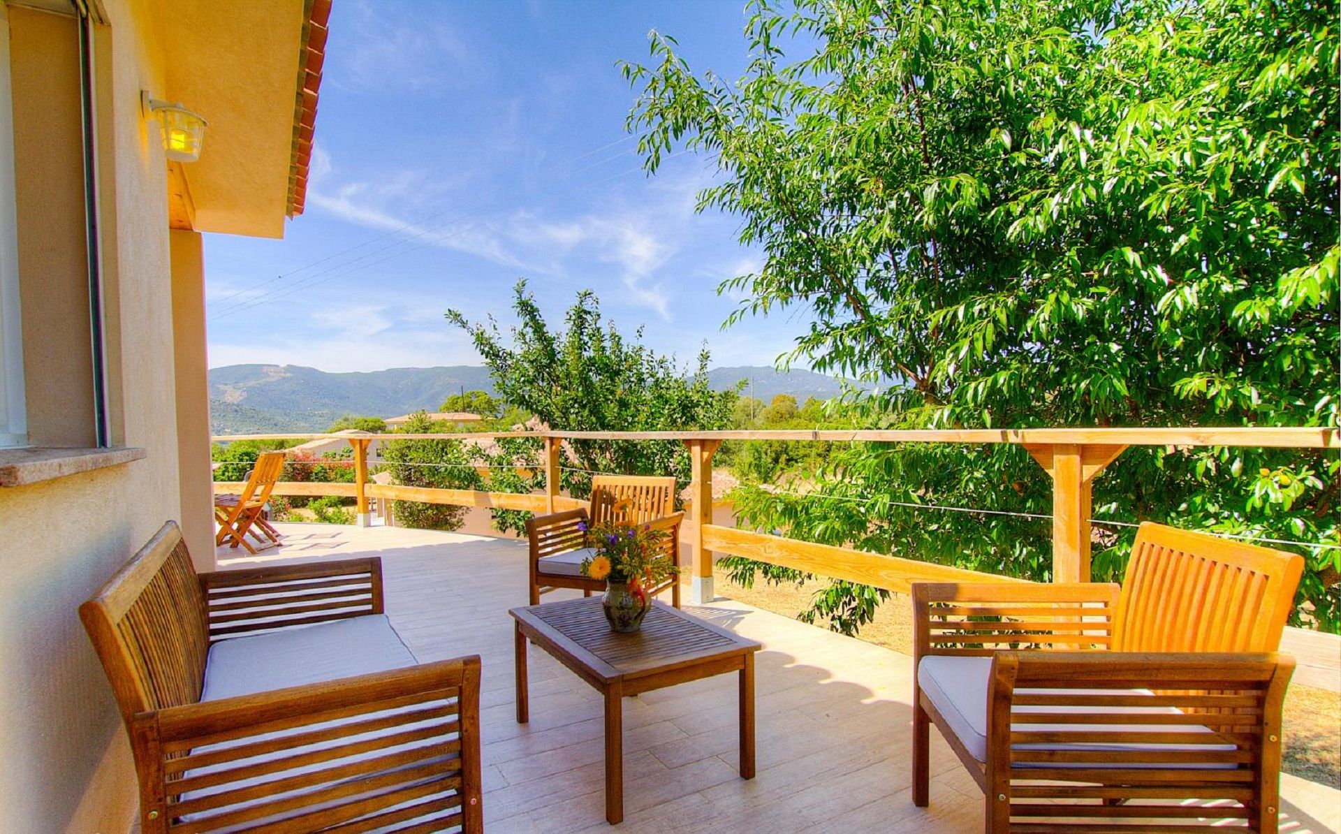 A wooden deck with outdoor furniture, a wooden railing, and a view of green trees and distant hills.