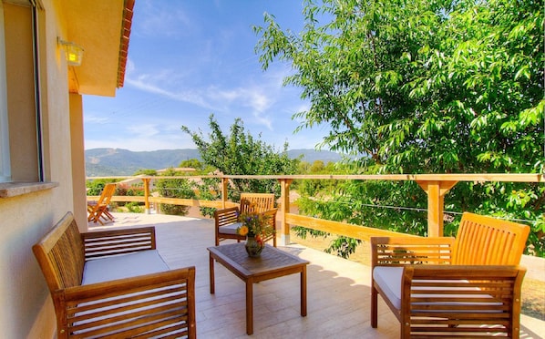 A wooden deck with outdoor furniture, a wooden railing, and a view of green trees and distant hills.