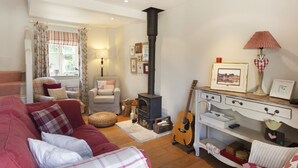 Un salon chaleureux avec une cheminée, un canapé, un fauteuil, une guitare et un bureau avec une lampe.