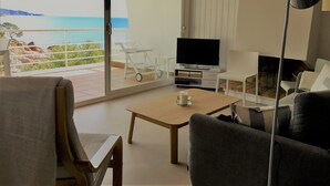 Ein modernes Wohnzimmer mit Fernseher, Couchtisch und Sitzgelegenheiten. Durch die Glastür ist ein Balkon mit Meerblick zu sehen.