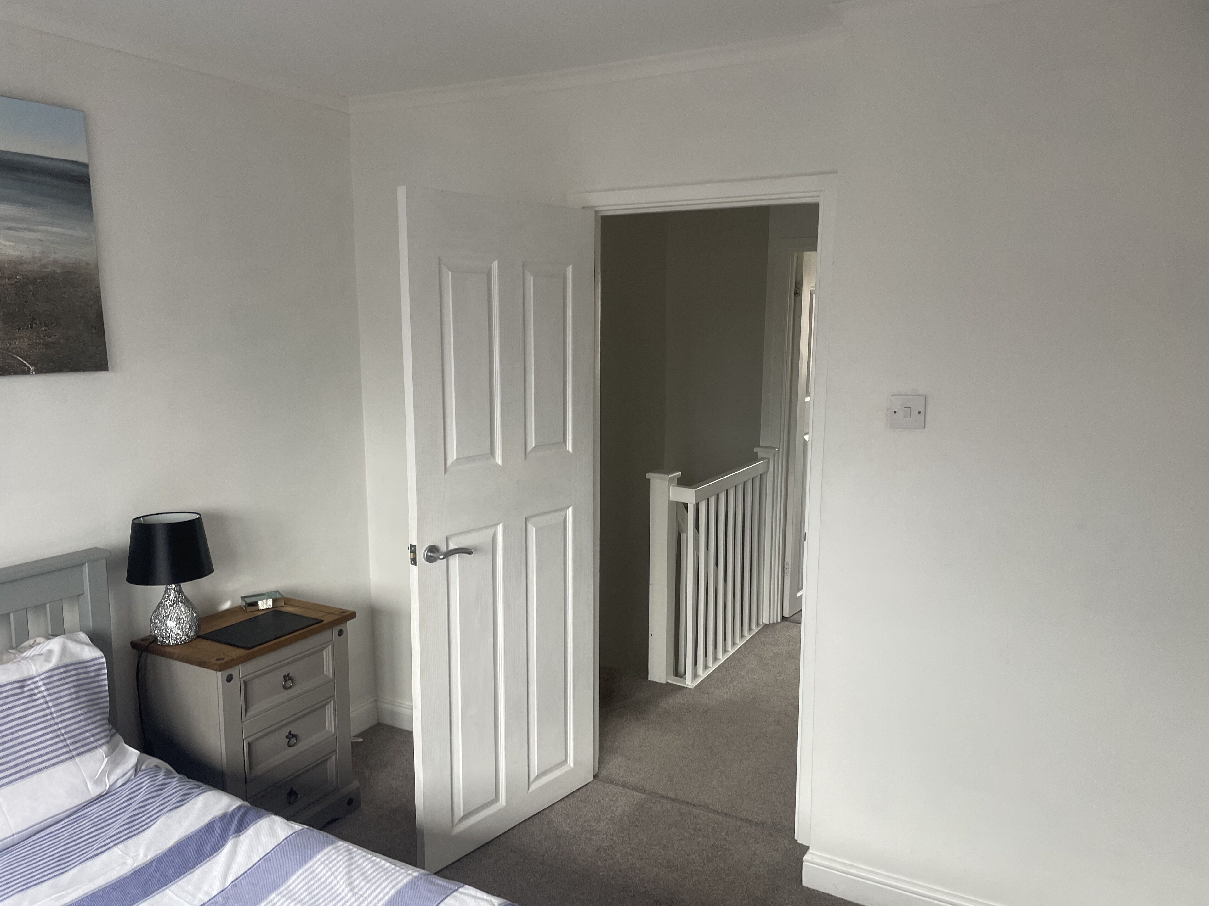 Une chambre à coucher avec un lit, une table de chevet, une lampe, une porte donnant sur un couloir et un tableau accroché au mur.