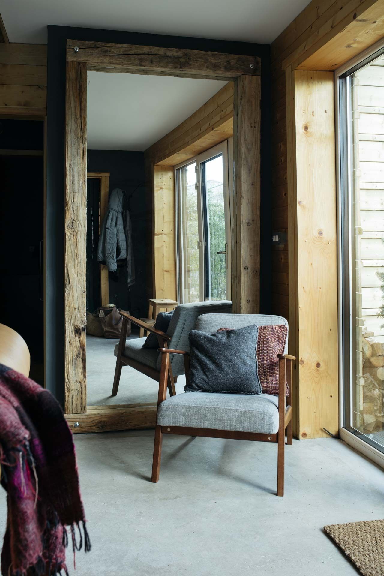 A cozy room with a wooden-framed window, a grey sofa with a patterned throw, and a large mirror reflecting the room.