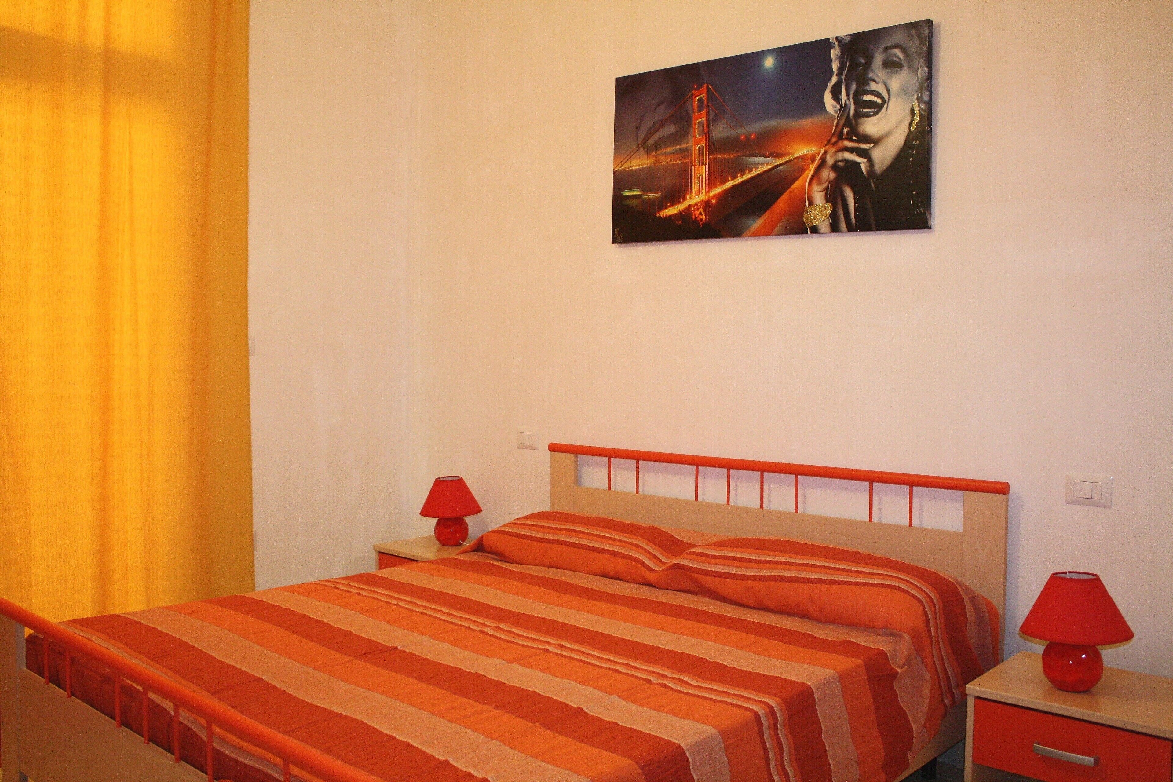 Une chambre à coucher avec un lit, deux tables de chevet, une commode, une lampe rouge et un tableau représentant un pont et une femme, accroché au mur.