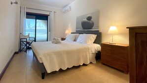 Quarto de hotel com cama, duas luminárias de cabeceira, uma mesinha de cabeceira de madeira, uma cadeira e vista para o mar através de uma porta deslizante.