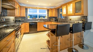 A modern kitchen with wooden cabinets, a large island with bar stools, a gas stove, and a view through the windows. - Forestside House - spacious house perfect for groups and families (Ballycastle)