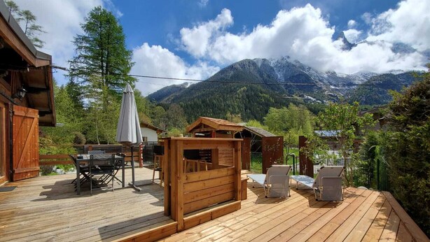 Um deck de madeira com área de estar ao ar livre, mesa e churrasqueira, com vista para uma paisagem montanhosa.