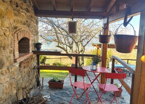 Een stenen muur, een bakoven, rode opklapbare stoelen, een ronde tafel, potplanten en een uitzicht op bomen en bergen.
