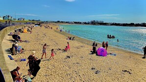 Una platja amb gent que pren el sol i nedant, una torre de socorristes i cases al fons.